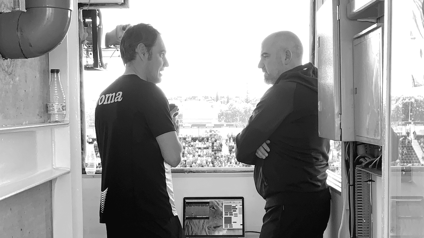 Two coaches talking in a stadium booth overlooking the pitch.